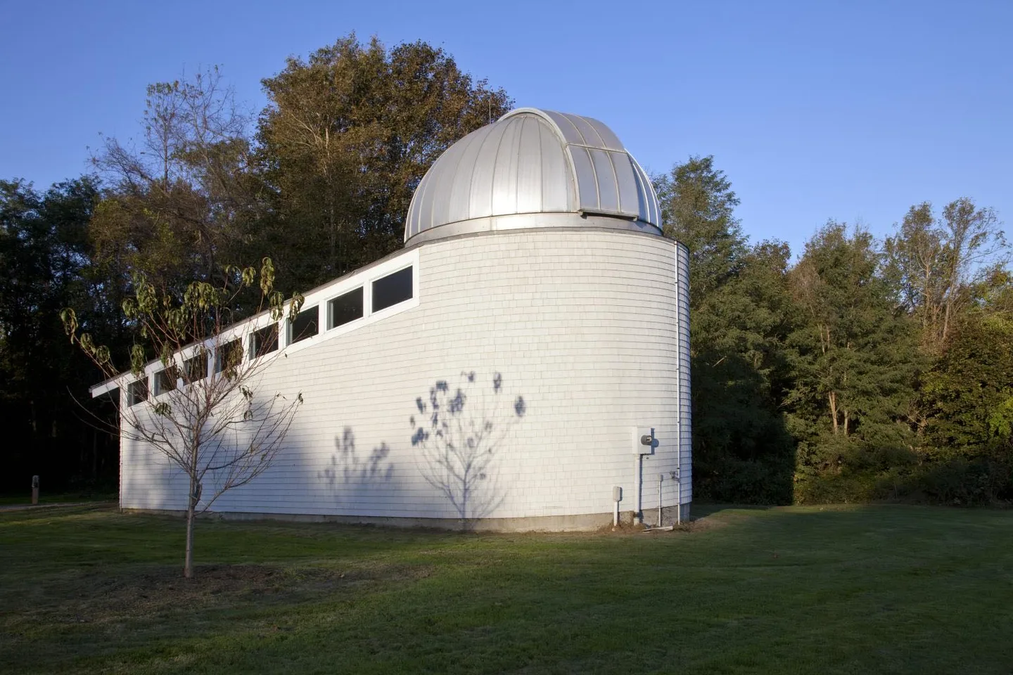 Color photograph of the white-shingled Alumni House Observatory with its silver dome, on a mowed lawn against trees and a clear blue sky. No people visible.