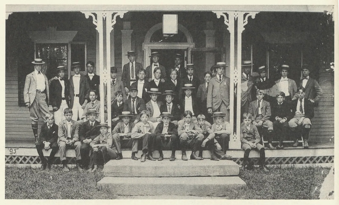 About three dozen boys in dark jackets, ties, and straw boater hats posed in rows on the ornate wooden porch and stone steps of a clapboard Victorian building, the original Pomfret Main House, in a faded black-and-white photograph.